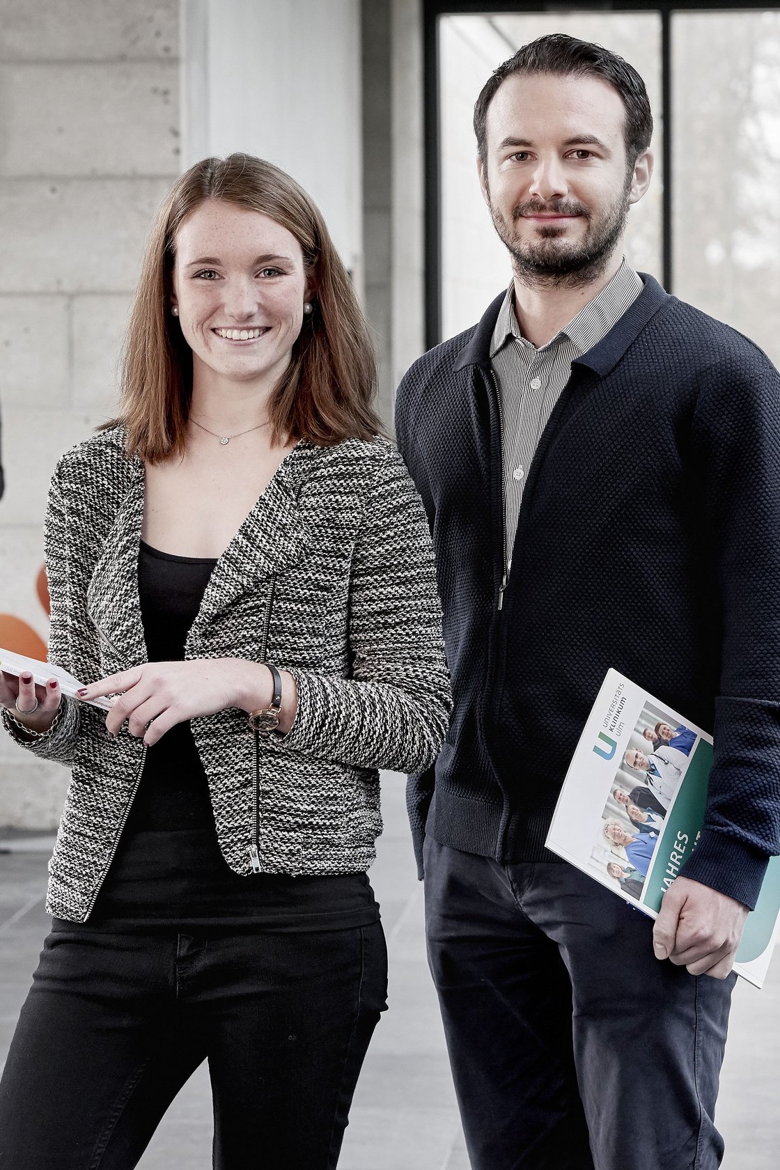 Zwei Trainees in Verwaltungsgebäude der Uniklinik vor einer Glasfront.